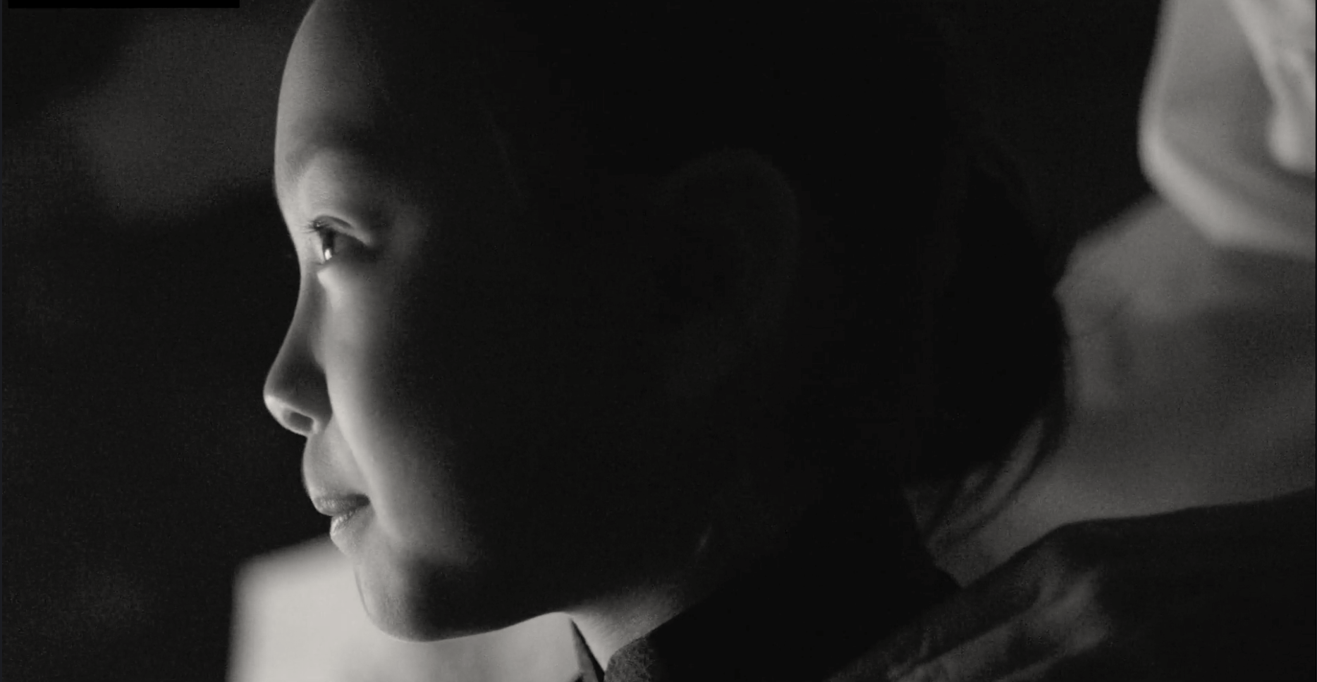 Person wearing a blouse with sheer sleeves, vest and skirt with back turned, reaching to close window shutters, silhouetted against the sunlit window, as a child looks on