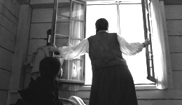 Person wearing a blouse with sheer sleeves, vest and skirt with back turned, reaching to close window shutters, silhouetted against the sunlit window, as a child looks on