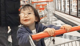 Young child with bandage over one eye, wearing glasses, smiling up at someone off-camera, standing in a supermarket aisle lined with freezer doors, pushing a cart, with the legs and coat of an adult visible behind the child.
