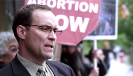 Man wearing glasses, blazer, shirt and tie with a stern expression among a crowd of people holding up signs. A partial sign is visible behind his head, red in the shape of a stop sign, with white block text 