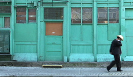 Side view of a person wearing black and a light-coloured brimmed hat walking past a green-painted building with a row of windows.